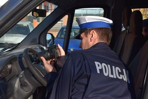 Zdjęcie w kolorze przedstawia stanowisko do przyjęcia pojazdów obsługiwane przez policjanta, który fotografuje pojazd typu bus.