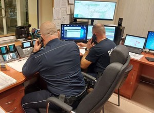 Zdjęcie w kolorze przedstawia pomieszczenie służby dyżurnej gdzie dwóch policjantów siedzi przy pulpicie z ekranami monitorów z telefonami przy uchu.