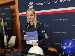 Zdjęcie w kolorze przedstawia stoisko policyjne z funkcjonariuszka pozuje do zdjęcia trzymając w dłoniach broszurę informacyjną.
