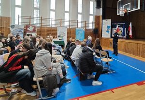 Zdjęcie w kolorze przedstawia policjanta z mikrofonem w dłoni stojącego po środku sali gimnastycznej, jest zwrócony twarzą do siedzących na krzesłach uczniów.
