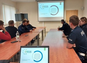 Zdjęcie w kolorze przedstawia na pierwszym planie ekran laptopa na nim wyświetlony slajd zawierający napis : równość płci w Policji. po lewej i prawej policjanci patrzący na ekran naścienny.