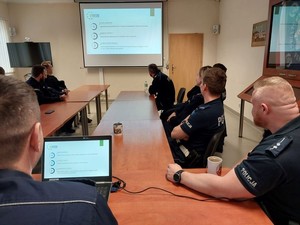 Zdjęcie w kolorze przedstawia na pierwszym planie ekran laptopa na nim wyświetlony slajd zawierający napis : równość płci w Policji. po lewej i prawej policjanci patrzący na ekran naścienny.
