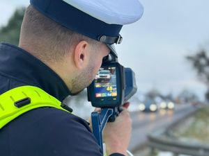 Zdjęcie w kolorze przedstawia policjanta z miernikiem prędkości skierowanym w stronę jadących pojazdów.