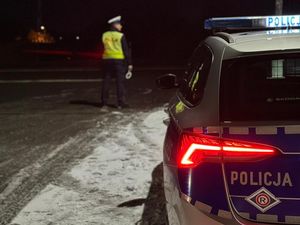 Zdjęcie w kolorze przedstawia porę wieczorowo - nocną, policjanta stojącego na jezdni z sygnalizatorem do zatrzymywania pojazdów, na pierwszym planie po prawej fragment radiowozu.