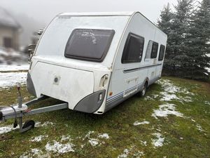 Zdjęcie w kolorze przedstawia przyczepę kempingową zaparkowaną w ogrodzie na podłożu trawiastym, w tle choinki.