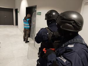 Zdjęcie w kolorze przedstawia policjantów w hełmach idących w stronę policjanta zespołu antykonfliktowego stojącego przy drzwiach.