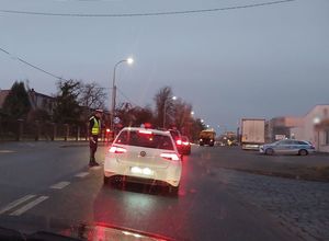 Zdjęcie w kolorze przedstawia policjanta z świecącym na czerwono sygnalizatorem prowadzi kontrolę trzeźwości kierującego pojazdem osobowym.