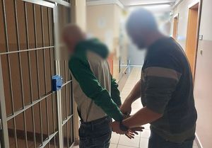 Zdjęcie w kolorze przedstawia policjanta , który stoi za mężczyzną który ma kajdanki założone na ręce trzymane z tyłu.