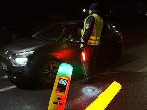 Zdjęcie w kolorze przedstawia porę nocną, na pierwszym planie sygnalizator do zatrzymywania pojazdów świecący na czerwono oraz analizator wydechu z zieloną lampką, na drugim planie sylwetka policjanta kontrolującego trzeźwość kierowcy samochodu osobowego.