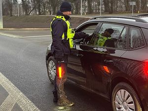 Zdjęcie w kolorze przedstawia dzień, na pierwszym planie policjant trzyma w ręku sygnalizator do zatrzymywania pojazdów świecący na czerwono oraz analizator wydechu - prowadzi  kontrolę trzeźwości kierowcy samochodem osobowym.