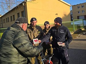 Zdjęcie w kolorze przedstawia policjanta w towarzystwie uczniów klasy mundurowej wręczających kartę seniorowi w tle żółty budynek.