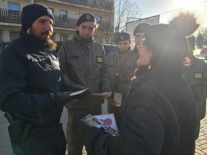 Zdjęcie w kolorze przedstawia policjanta w towarzystwie uczniów klasy mundurowej wręczających kartę seniorce.