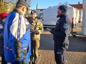 Zdjęcie w kolorze przedstawia policjanta w towarzystwie uczniów klasy mundurowej wręczających kartę seniorom w tle zabudowania.