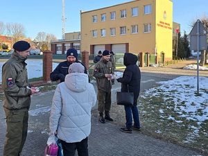 Zdjęcie w kolorze przedstawia policjanta w towarzystwie uczniów klasy mundurowej wręczających kartę seniorom w tle żółty budynek.