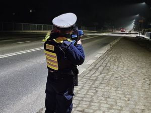 Zdjęcie w kolorze przedstawia policjantkę ruchu drogowego w nocnej scenerii mierzy prędkość odjeżdżającego pojazdu.