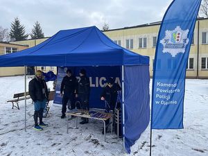 Zdjęcie w kolorze przedstawia niebieski namiot z napisem policja, przy nim kilka osób oraz policjanci, do koła śnieg.