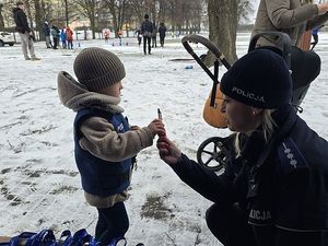 Zdjęcie w kolorze przedstawia policjantkę wręczającą małemu chłopcu element odblaskowy. W tle biegające po zaśnieżonej bieżni osoby.