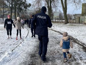 Zdjęcie w kolorze przedstawia na pierwszym planie policjanta biegnącego z małym chłopcem, dalej pozostałe osoby uczestniczące w biegu.
