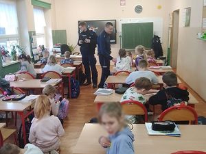 zdjęcie w kolorze przedstawia policjantów stojących pomiędzy ławkami w klasie, dzieci siedzą w ławkach.
