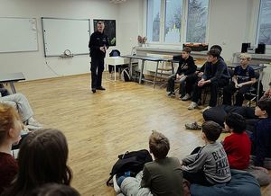 Zdjęcie w kolorze przedstawia policjanta podczas prelekcji w szkole, dzieci i młodzież siedzą do koła na krzesłach policjant stoi po środku.