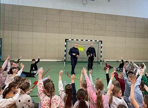 Zdjęcie w kolorze przedstawia policjantów podczas prelekcji w szkole na sali gimnastycznej, dzieci i młodzież siedzą do koła na podłodze policjanci stoją po środku, za nimi bramka piłkarska.