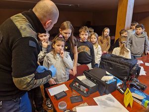 Zdjęcie w kolorze przedstawia dzieci zgromadzone przy stole, na którym poukładane są różne elementy wyposażenia policyjnego Technika Kryminalistyki - funkcjonariusz stoi obok grupy dzieci.