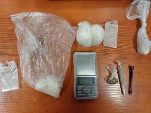 Zdjęcie w kolorze, przedstawia worki foliowe z białym proszkiem oraz mały woreczek foliowy z suszem roślinnym, obok fajka szklana do papierosów oraz waga jubilerska. Wszystkie przedmioty położone na drewnianym blacie.