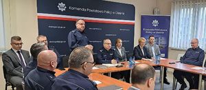 Świetlica Komendy Powiatowej Policji w Oleśnie. Przy stołach ustawionych w podkowę siedzą umundurowani policjanci oraz zaproszeni goście. Przy stole na tle granatowego baneru z napisem Komenda Powiatowa Policji w Oleśnie stoi Komendant Powiatowy Policji w Oleśnie inspektor Rafał Drozdowski.