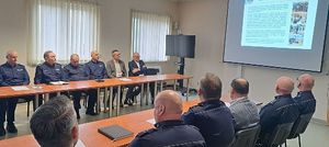 Świetlica Komendy Powiatowej Policji w Oleśnie. Przy stołach ustawionych po dwóch stronach wzdłuż ścian siedzą umundurowani policjanci i patrzą na ekran zawieszony na ścianie gdzie wyświetlany jest kolorowy slajd prezentacji. Treść slajdu nieczytelna.