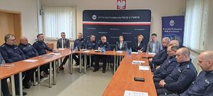 Świetlica Komendy Powiatowej Policji w Oleśnie. Przy stołach ustawionych w podkowę siedzą policjanci w mundurach służbowych oraz zaproszeni goście. W tle granatowy baner z napisem Komenda Powiatowa Policji w Oleśnie.