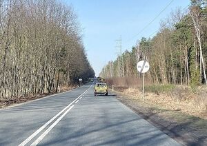 Zdjęcie w kolorze przedstawia drogę w obszarze leśnym, na jezdni pojazdy jadące w oddali na przeciwnych pasach ruchu.