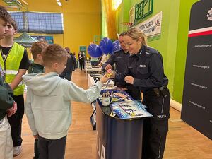 Zdjęcie w kolorze przedstawia policyjne stanowisko promocyjne ze ścianką z płótna w kolorze granatowym. Policjantka stoi za blatem z napisem policja po lewej stronie młodzi chłopcy odbierają od policjantki elementy promocyjne.