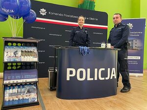 Zdjęcie w kolorze przedstawia policyjne stanowisko promocyjne ze ścianką z płótna w kolorze granatowym i blatem z białym napisem policja granatowym tle . Policjantka stoi za blatem, obok po prawej policjant . Za jego plecami plakat rozwijany z rolki z napisem Komenda Powiatowa Policji w Oleśnie.