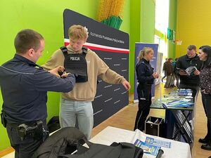 Zdjęcie w kolorze przedstawia policyjne stanowisko promocyjne ze ścianką z płótna w kolorze granatowym. Policjantka stoi za blatem z napisem policja po lewej stronie policjant pomaga założyć młodemu mężczyźnie kamizelkę  kuloodporną z napisem policja.