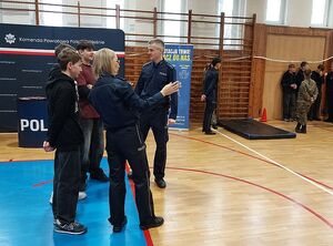 Zdjęcie w kolorze przedstawia uczniów szkół  z policjantkami, w tle sala gimnastyczna, policjantka pokazuje palcem kierunek biegu.