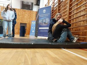 Zdjęcie w kolorze przedstawia młoda kobietę wykonująca skłony z leżenia na materacu w asyście policjantki.