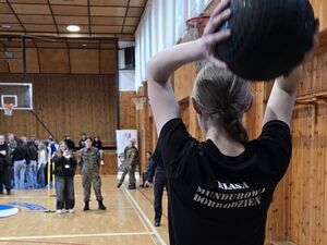 Zdjecie w kolorze przedstawia uczennicę w koszulce z napisem klasa mundurowa, która trzyma piłke nad głowa gotowa do rzutu.