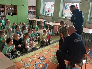 Zdjęcie w kolorze przedstawia przedszkolaków siedzących na kolorowym dywanie, na pierwszym planie  dwaj policjanci jeden stoi a drugi siedzi na małym krześle obok nich stoi jeden z przedszkolaków