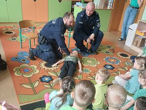 Zdjęcie w kolorze przedstawia przedszkolaków siedzących na kolorowym dywanie, na środku dwaj policjanci w pozycji kucznej przy leżacym na dywanie chłopcu.