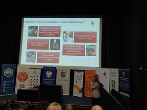 Zdjęcie w kolorze przedstawia slajd na temat bezpieczeństwa bankowego wyświetlony w sali kinowej, poniżej slajdu liczne plakaty rozwijane z rolek zawierające nazwy partnerów konferencji, w dolnym prawym rogu kobieta z mikrofonem w ręku.