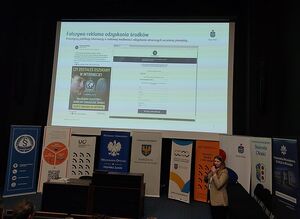 Zdjęcie w kolorze przedstawia slajd na temat bezpieczeństwa bankowego wyświetlony w sali kinowej, poniżej slajdu liczne plakaty rozwijane z rolek zawierające nazwy partnerów konferencji, w dolnym prawym rogu kobieta z mikrofonem w ręku.