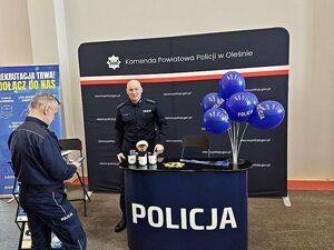 Zdjęcie w kolorze przedstawia stanowisko promocyjne i dwóch policjantów, jeden z nich stoi przodem a drugi bokiem .