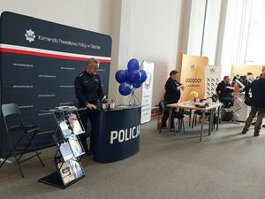 Zdjęcie w kolorze przedstawia stanowisko promocyjne i policjanta, w tle stanowiska innych firm i  instytucji.