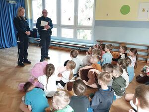 Zdjęcie w kolorze, przedstawia kilkudziesięciu  przedszkolaków  siedzących na parkiecie, spoglądają na parę policjantów stojących przed nimi.