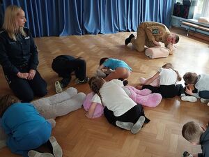 Zdjęcie w kolorze, przedstawia   przedszkolaków  ćwiczących czynności resuscytacji na maskotkach. Pokazu udzielają Strażak i policjantka.