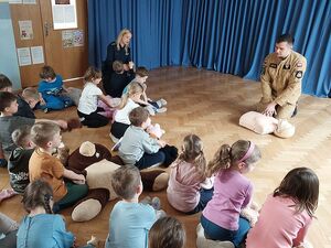 Zdjęcie w kolorze, przedstawia   przedszkolaków  ćwiczących czynności resuscytacji na maskotkach. Pokazu udzielają Strażak i policjantka.