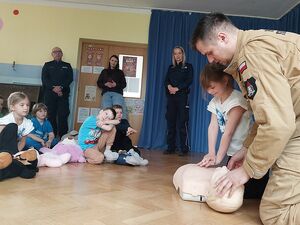 Zdjęcie w kolorze, przedstawia strażaka udzielającego instrukcji resuscytacji na leżacym przed nim manekinie, w tle  przedszkolaki oraz policjanci i opiekunki.