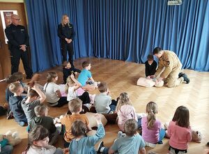Zdjęcie w kolorze, przedstawia   przedszkolaków  ćwiczących czynności resuscytacji na maskotkach. Pokazu udzielają Strażak na lezacym przed nim fantomie.