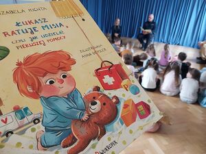 Zdjęcie w kolorze przedstawia fragment okładki książki pod tytułem Łukasz Ratuje Misia, w tle przedszkolaki i funkcjonariusz siedzacy na krześle czytający książeczkę.