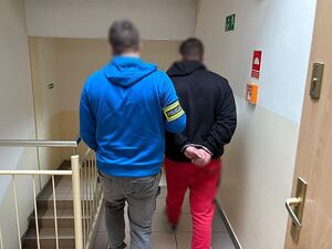 Zdjęcie w kolorze przedstawia dwóch mężczyzn, po lewej policjant w ubraniu cywilnym trzyma za rękę mężczyznę po prawej, który ma kajdanki na rękach trzymanych z tyłu.